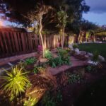 Jardín iluminado al anochecer con plantas diversas, flores moradas y un cercado de madera. La escena muestra un ambiente natural y acogedor, ideal para espacios exteriores.