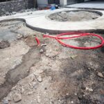 Terreno en construcción con área de concreto parcialmente terminada, mangueras rojas visibles y materiales de construcción esparcidos. Fondo de una casa moderna en proceso de edificación.