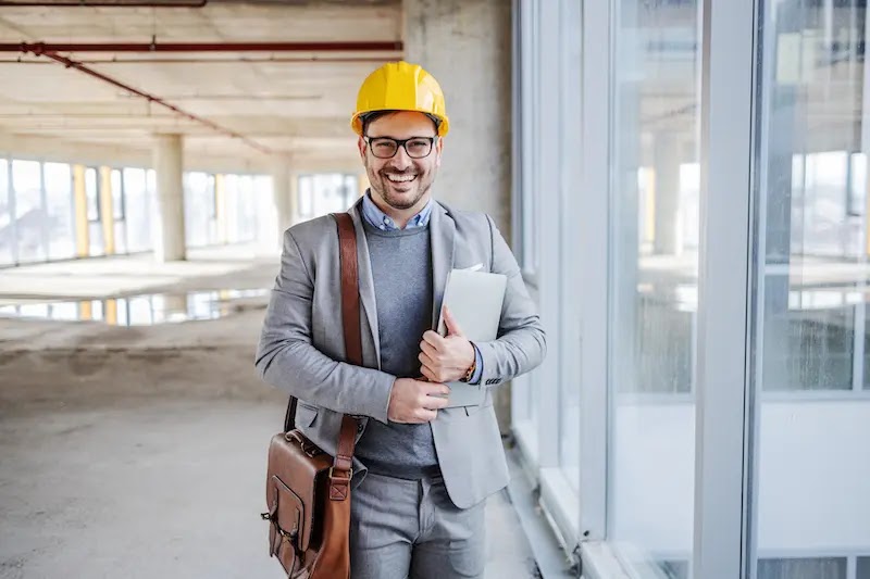 Hombre sonriente con casco de seguridad y traje, sosteniendo documentos en un sitio de construcción. Fondo con ventanas grandes y estructura de edificio en construcción.
